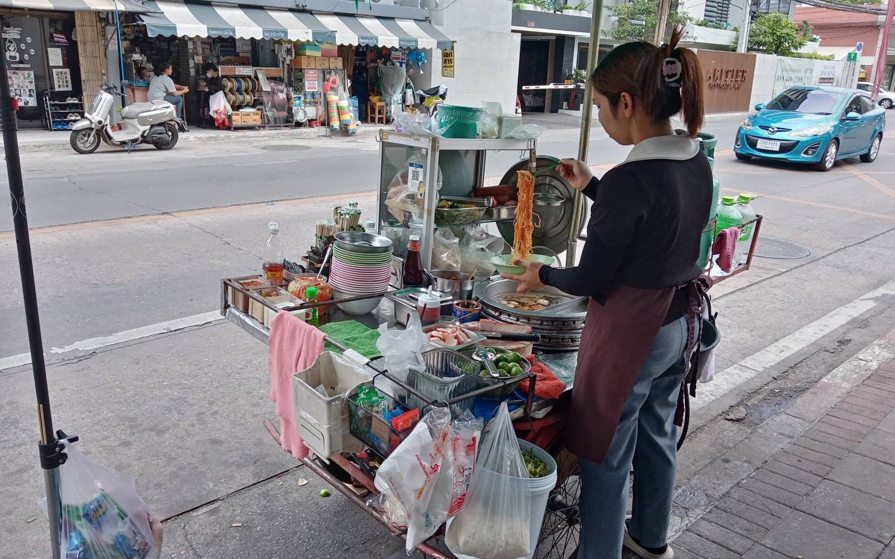 A cook portioning yellow noodles into a bowl at a street cart under a green umbrella