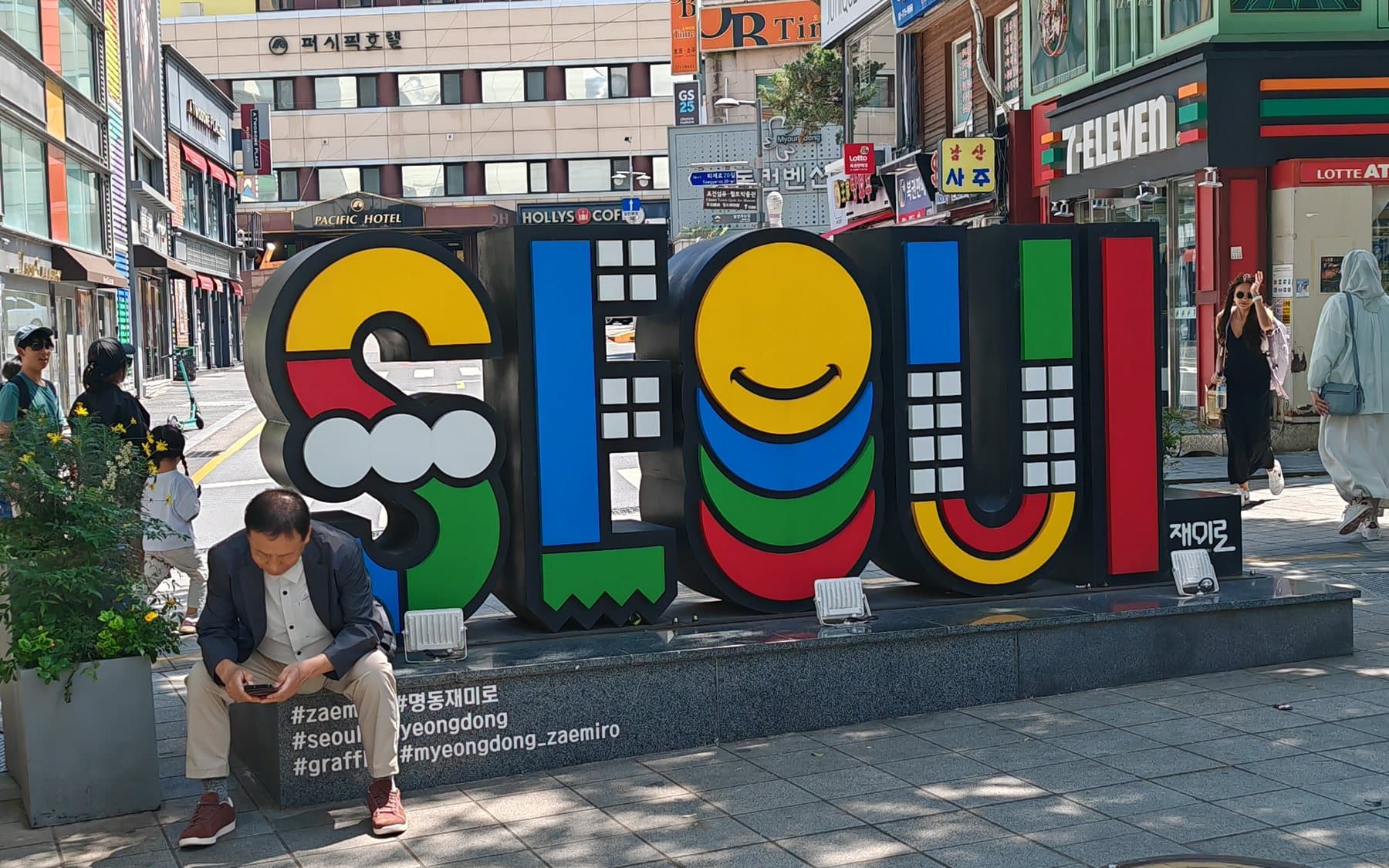 Colorful SEOUL sculpture on a Myeongdong sidewalk, with shopfronts and a few pedestrians on a sunny afternoon
