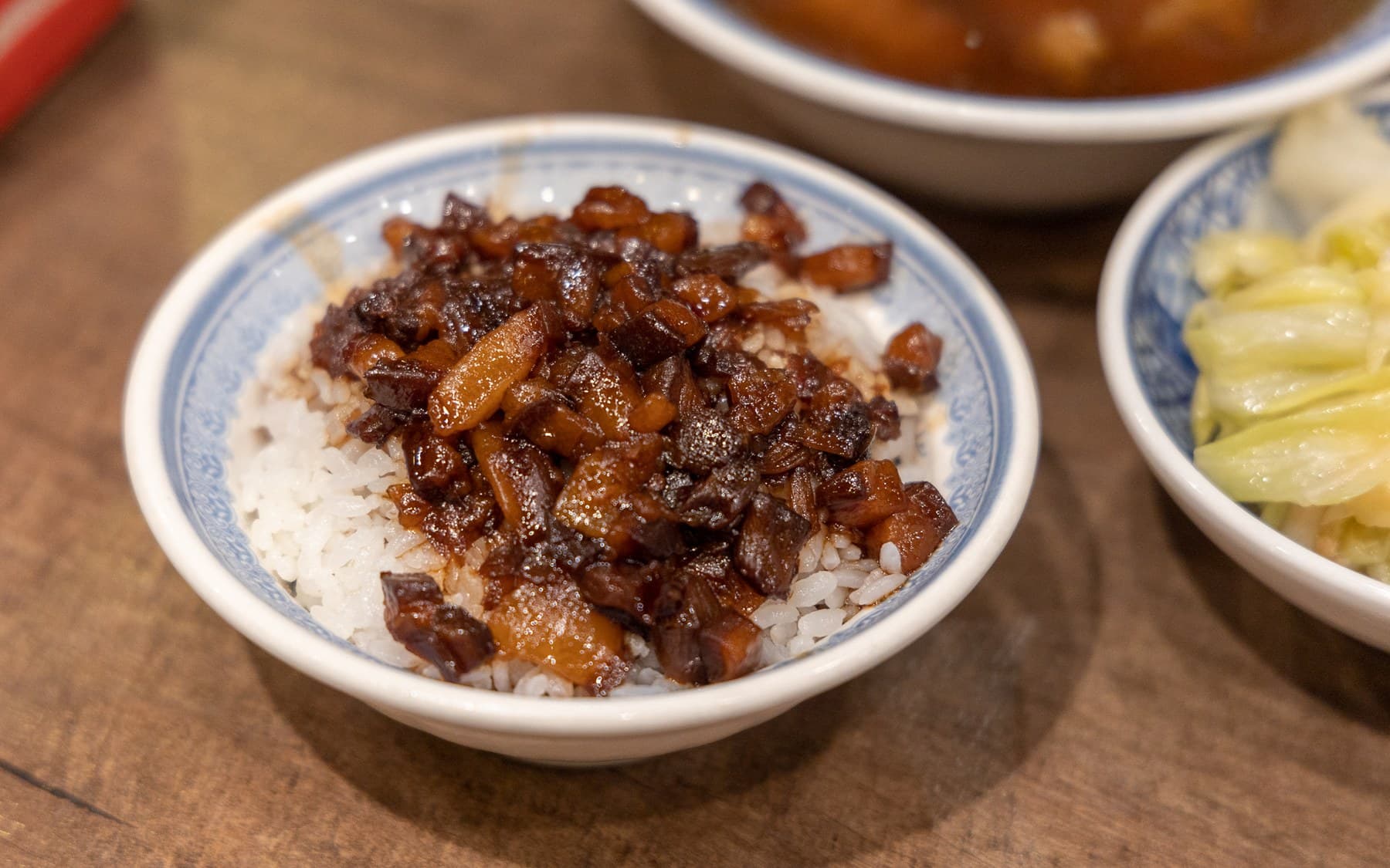 A small bowl of braised pork over white rice, with cabbage and soup at the edge of the table