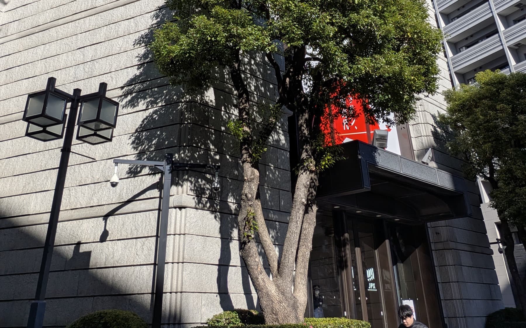Stone facade and entrance of the Tokyo Stock Exchange, with a tree in front and the JPX mark on the door